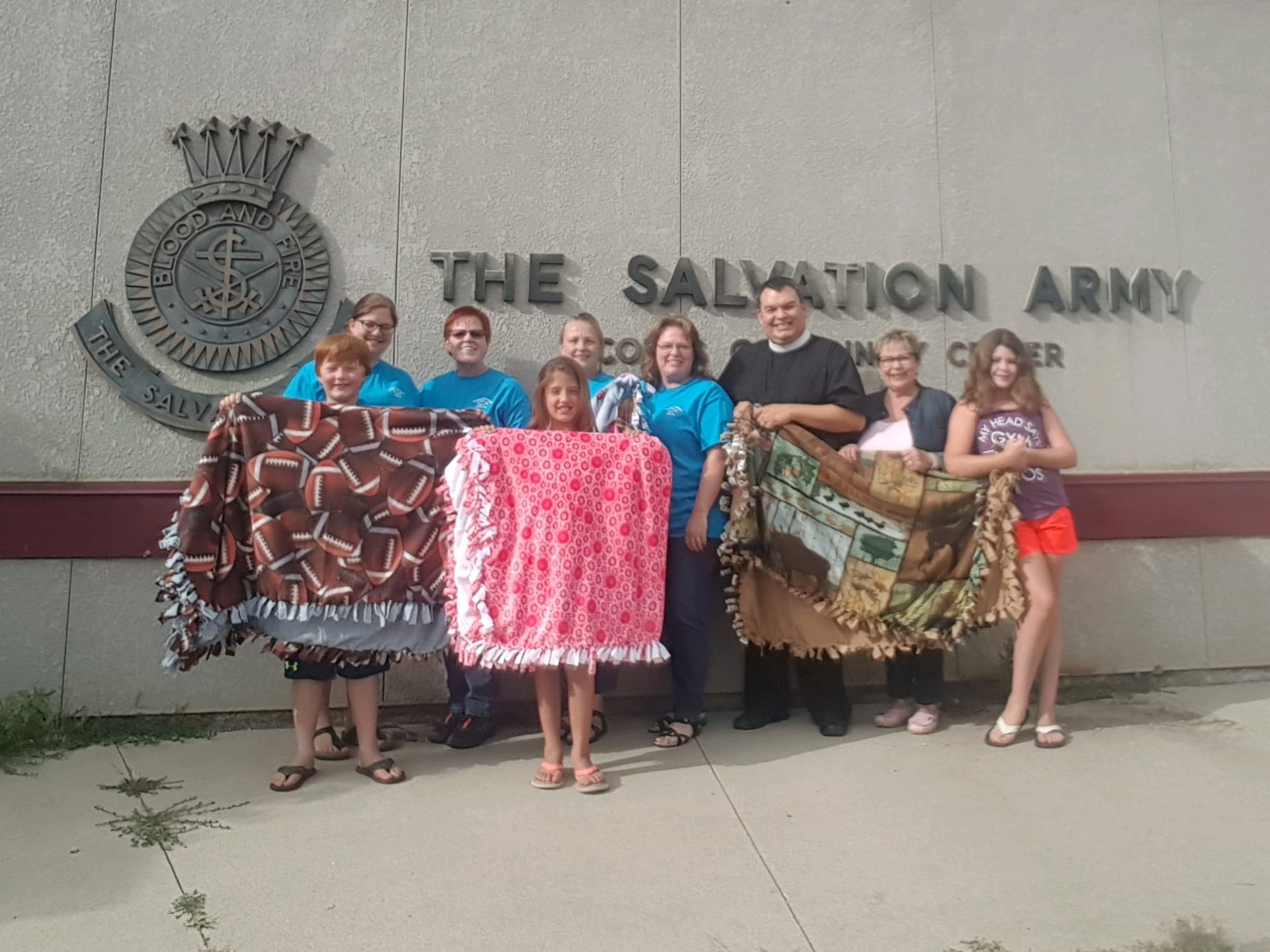 Nine people of various ages holding homemade tie blankets in front of a Salvation Army for donation